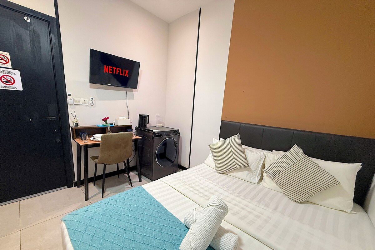 A hotel room with a bed, a desk with a computer, a washing machine, and a TV displaying the Netflix logo.