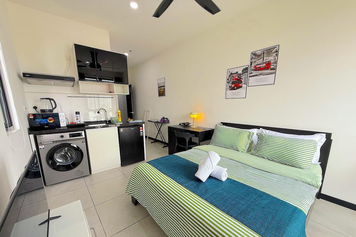 A compact hotel room with a bed, a small kitchenette, a washing machine, and a desk with a chair.