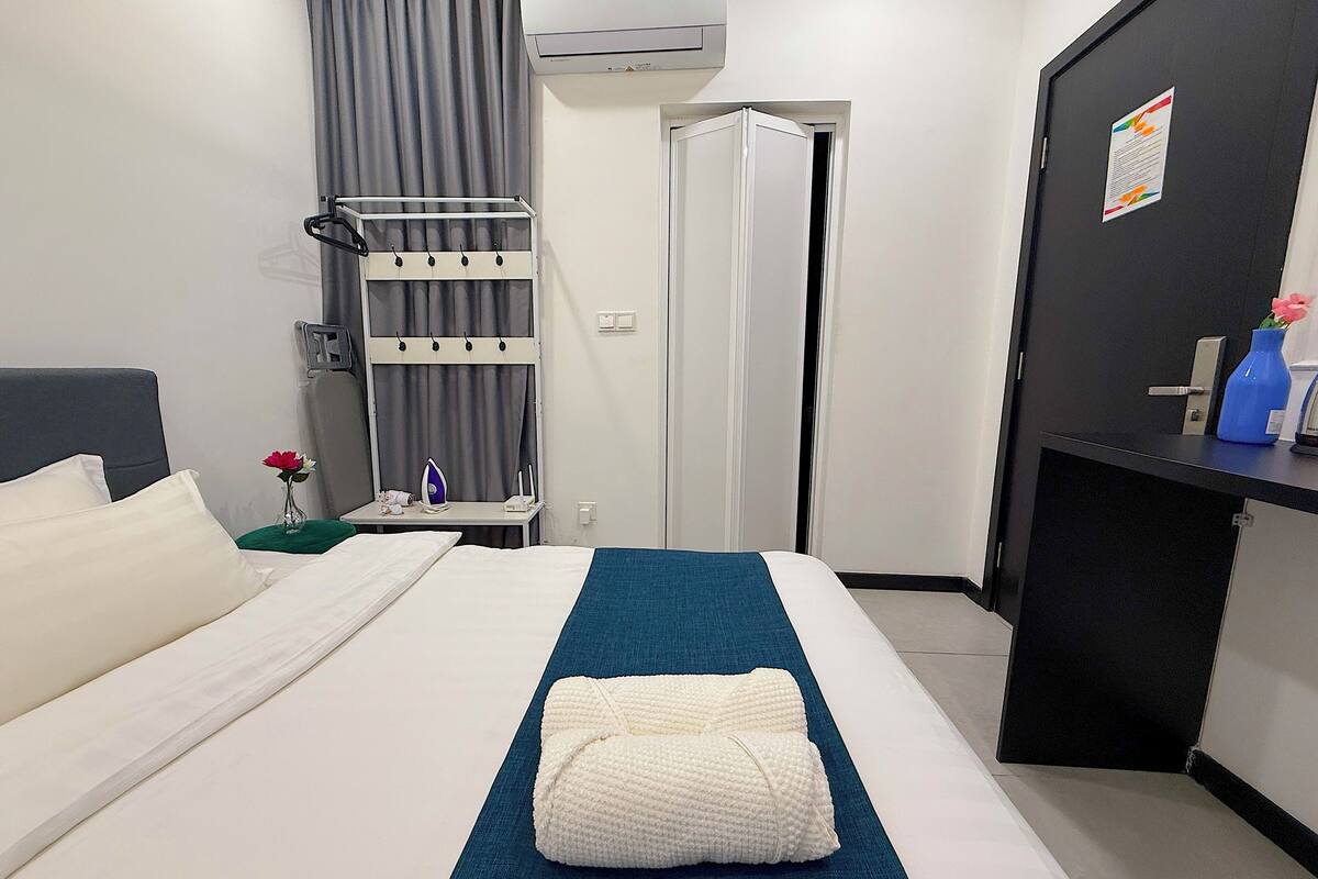 A hotel room with a bed, a desk with an ironing board, a wall-mounted air conditioner, and a door with a notice.