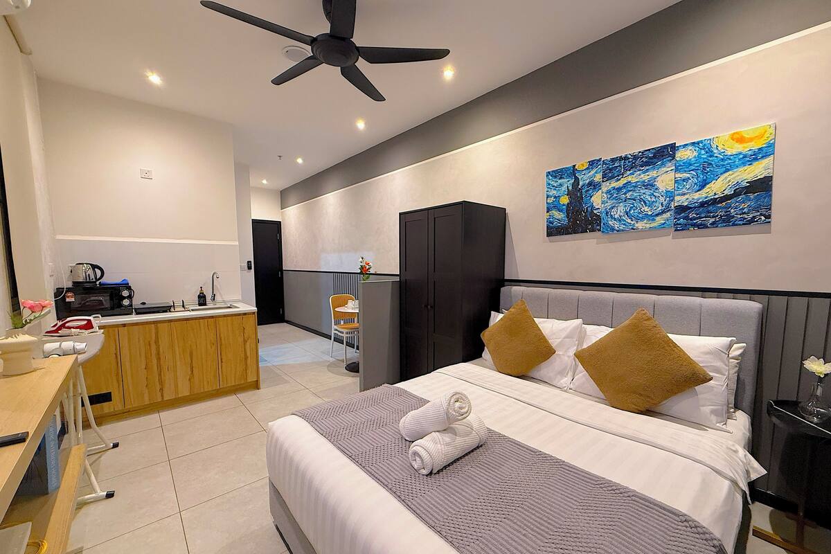 A modern hotel room with a bed, a desk, a kitchenette, and a ceiling fan.