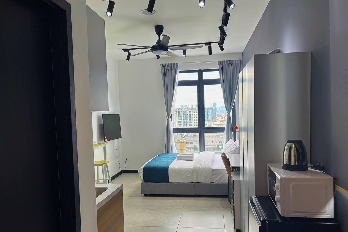 A modern hotel room with a bed, a desk, a TV, a ceiling fan, and a view of the city.