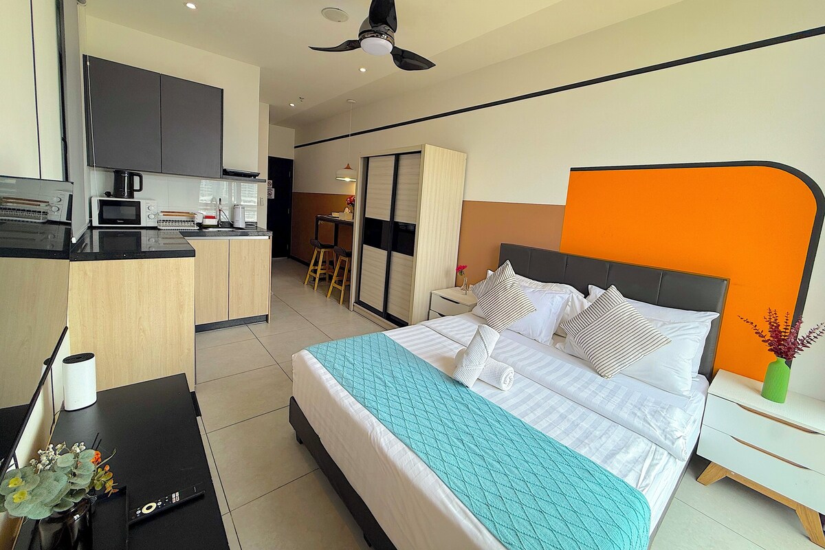 A modern hotel room with a bed, bedside table, and a kitchenette area.