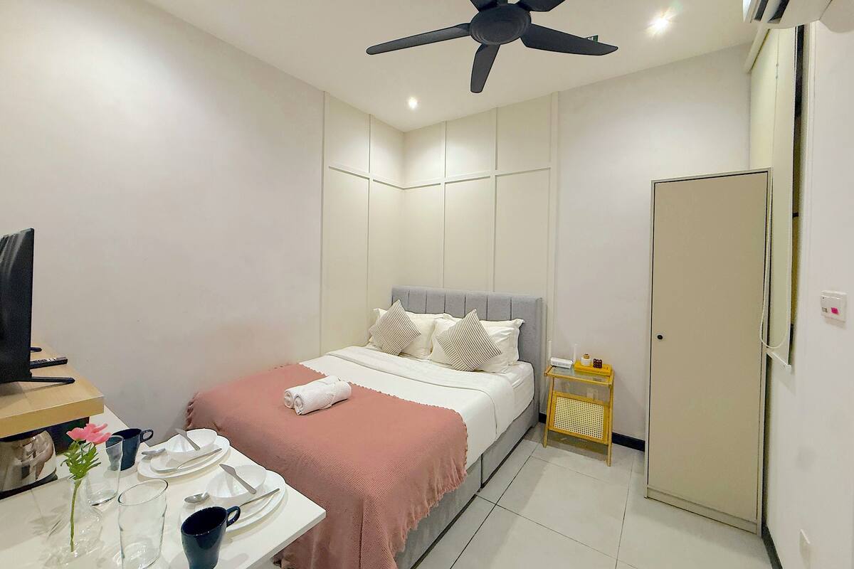A hotel room with a bed, a desk with a computer, a ceiling fan, and a door.