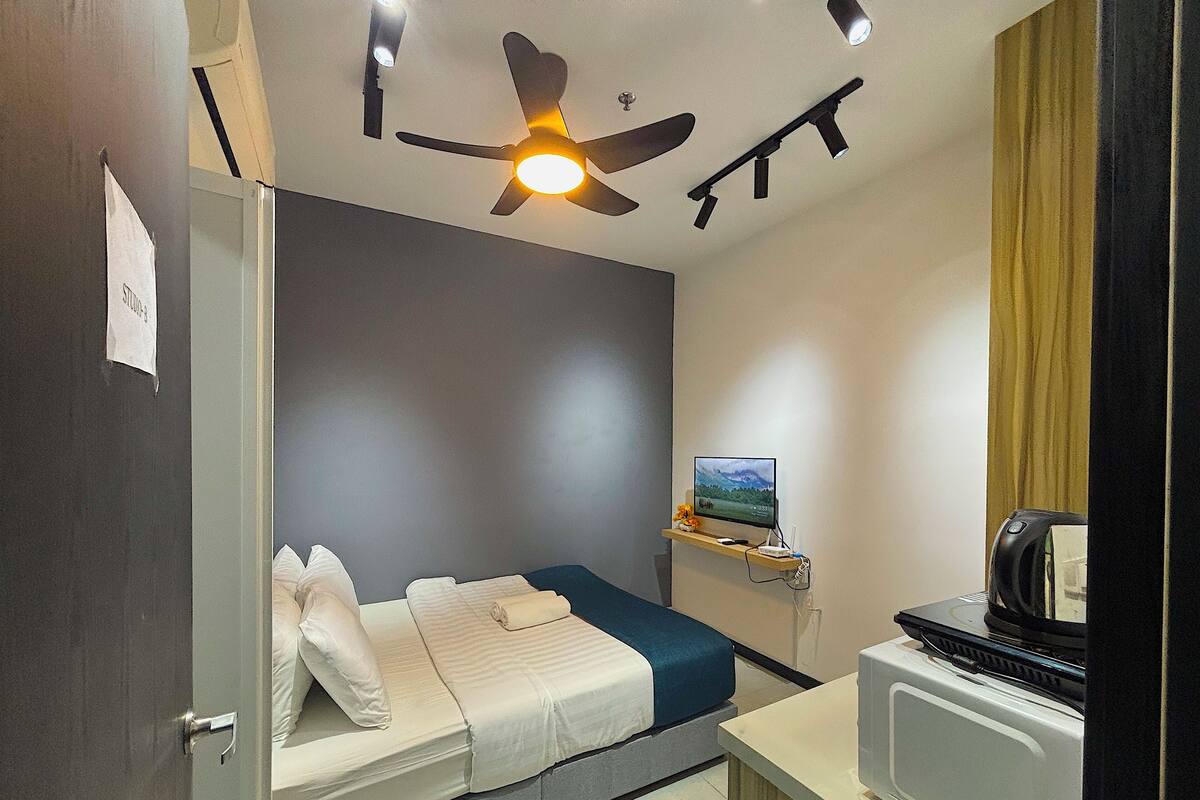 A compact hotel room with a single bed, a small desk with a computer, a ceiling fan, and a TV on a shelf.