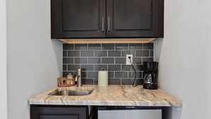 Una cocina compacta con armarios oscuros, encimera de mármol, un lavamanos, cafetera y refrigerador.