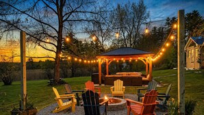 Ein Terrassenbereich im Freien mit Adirondack-Stühlen, einer Feuerstelle und einer überdachten Sitzgelegenheit unter Lichterketten.