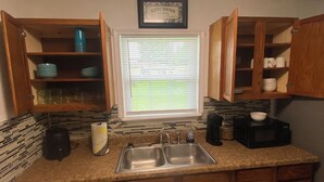 A compact kitchen with wooden cabinets, a double sink, a microwave, and a window with blinds. - “Cozy Diamond on the Corner” (Port Arthur)