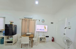 Una habitación pequeña y bien iluminada con una cama individual, un escritorio con una silla, un refrigerador negro, un televisor montado en la pared y un pequeño ventilador.