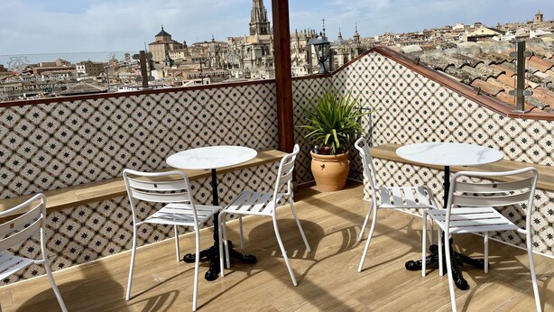 Um terraço com duas mesas e cadeiras brancas, um vaso de planta e uma parede com padrão.