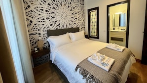 A neatly made bed with a white comforter and two pillows, a decorative wall panel, a bedside table with a lamp, and a mirror above the sink. - Casón De Los López Apartments 1B (Toledo)