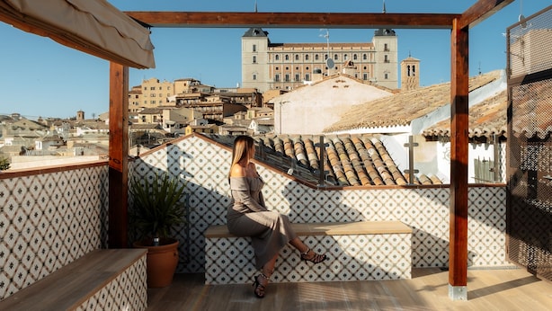 A balcony with patterned tiles, a potted plant, and a view of historic buildings in the background. - Casón De Los López Apartments 1B (Toledo)