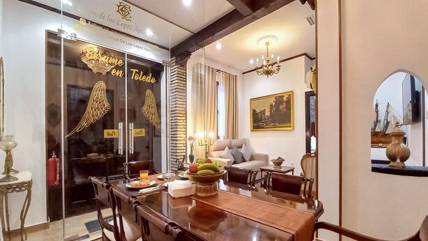 Uma sala de jantar com uma mesa de madeira, cadeiras e uma fruteira. Há um sofá e um lustre ao fundo.