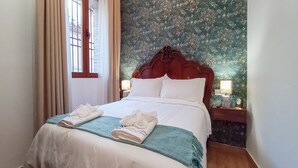 Cama bem arrumada com lençóis brancos e manta azul, duas toalhas brancas dobradas sobre a cama, cabeceira de madeira e mesa de cabeceira com abajur.