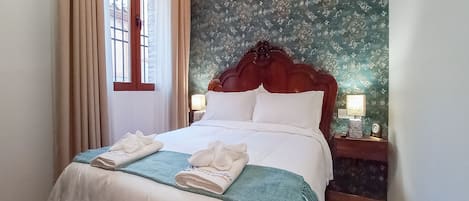 A neatly made bed with white linens and a blue throw blanket, two white towels folded on the bed, a wooden headboard, and a nightstand with a lamp.
