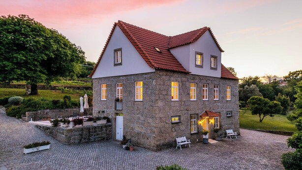 A stone building with a red-tiled roof, multiple windows, and a small outdoor seating area with benches and umbrellas. - Quinta Lusitânia (Santa Comba Dao)