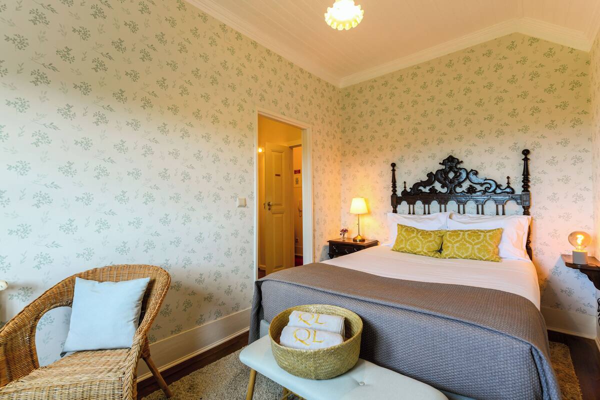A bedroom with floral wallpaper, a wicker chair, a bedside table with a lamp, and a bed with a headboard.