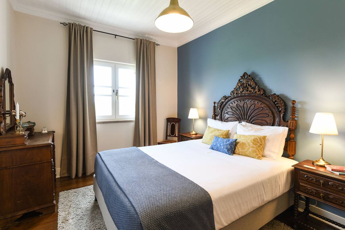 A bedroom with a large bed, wooden headboard, two bedside tables with lamps, a dresser, and a window with curtains.