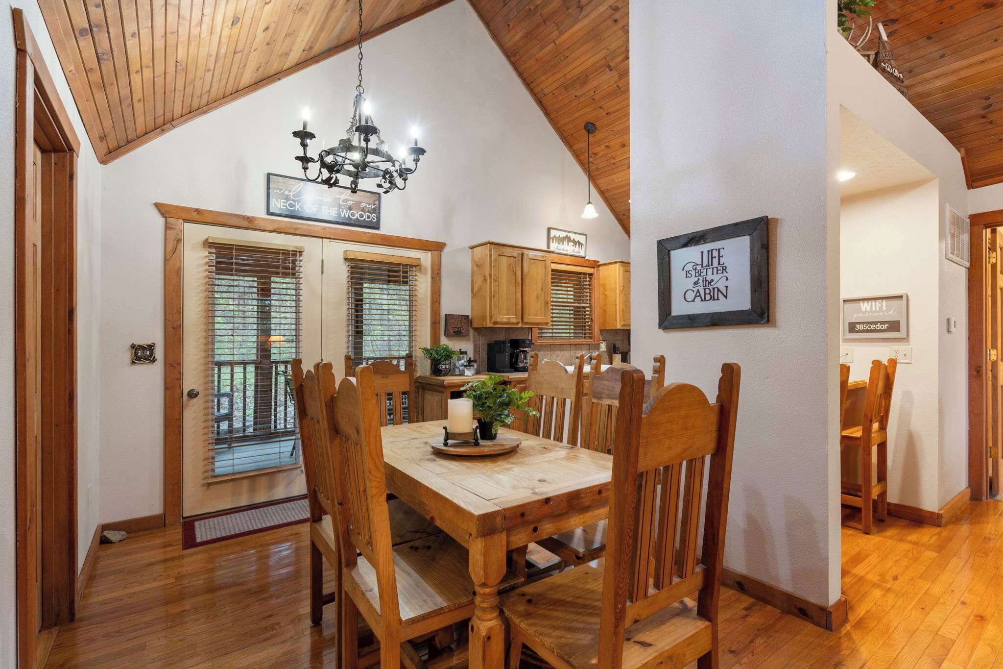 A dining area with a wooden table and chairs, a chandelier, and a kitchen in the background.