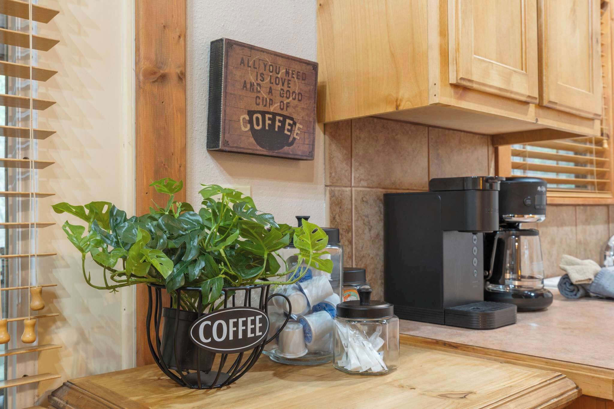 A kitchen with wooden cabinets, a coffee machine, a potted plant, and a sign that reads "All you need is love and a good cup of coffee."