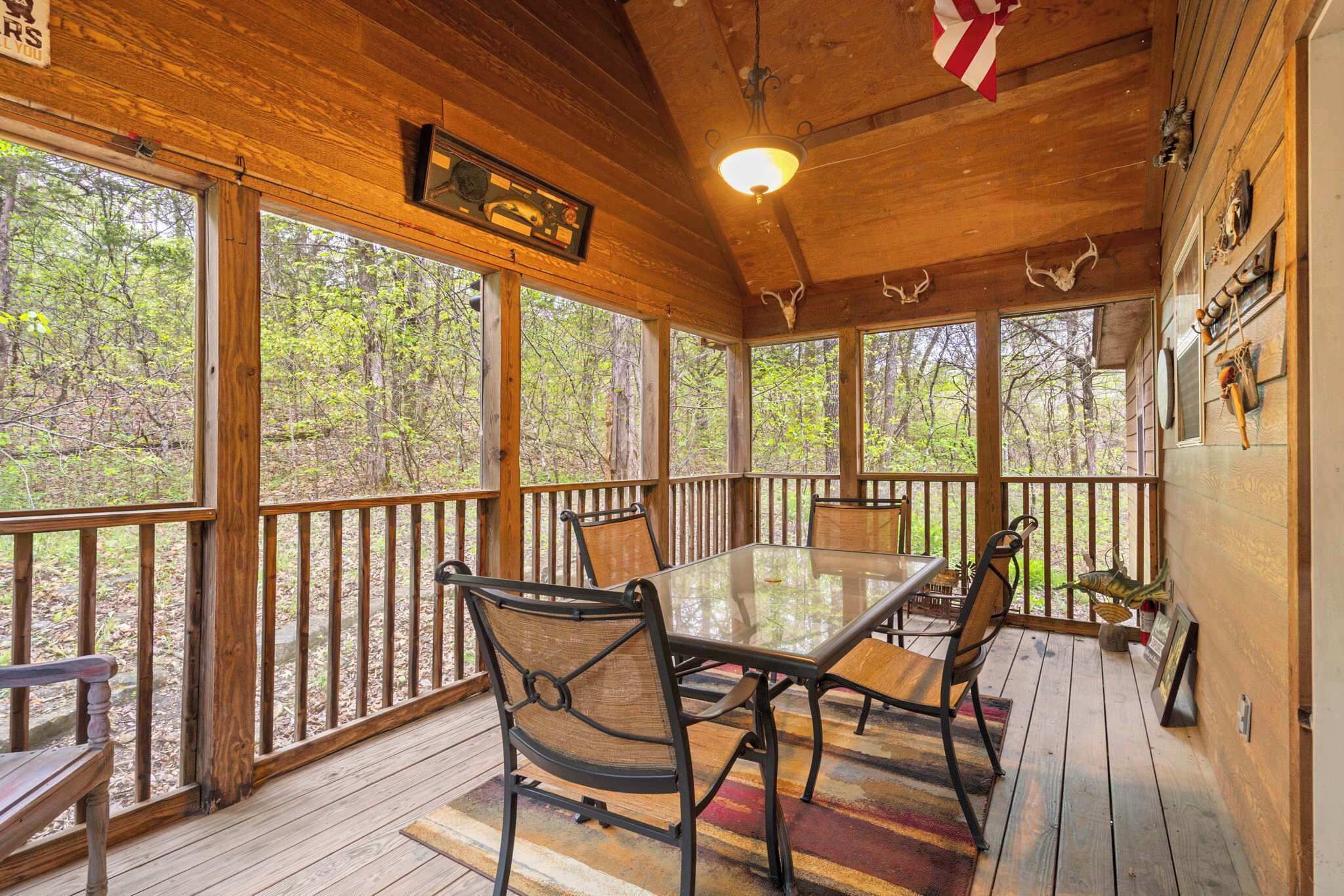 A wooden deck with a glass table and chairs, surrounded by large windows offering a forest view.