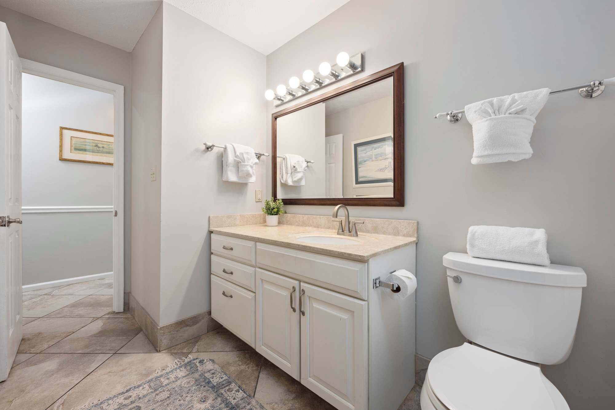 Une salle de bain avec un lavabo blanc, un meuble-lavabo et des toilettes. Plusieurs serviettes sont suspendues à des crochets et un tableau encadré est accroché au mur.