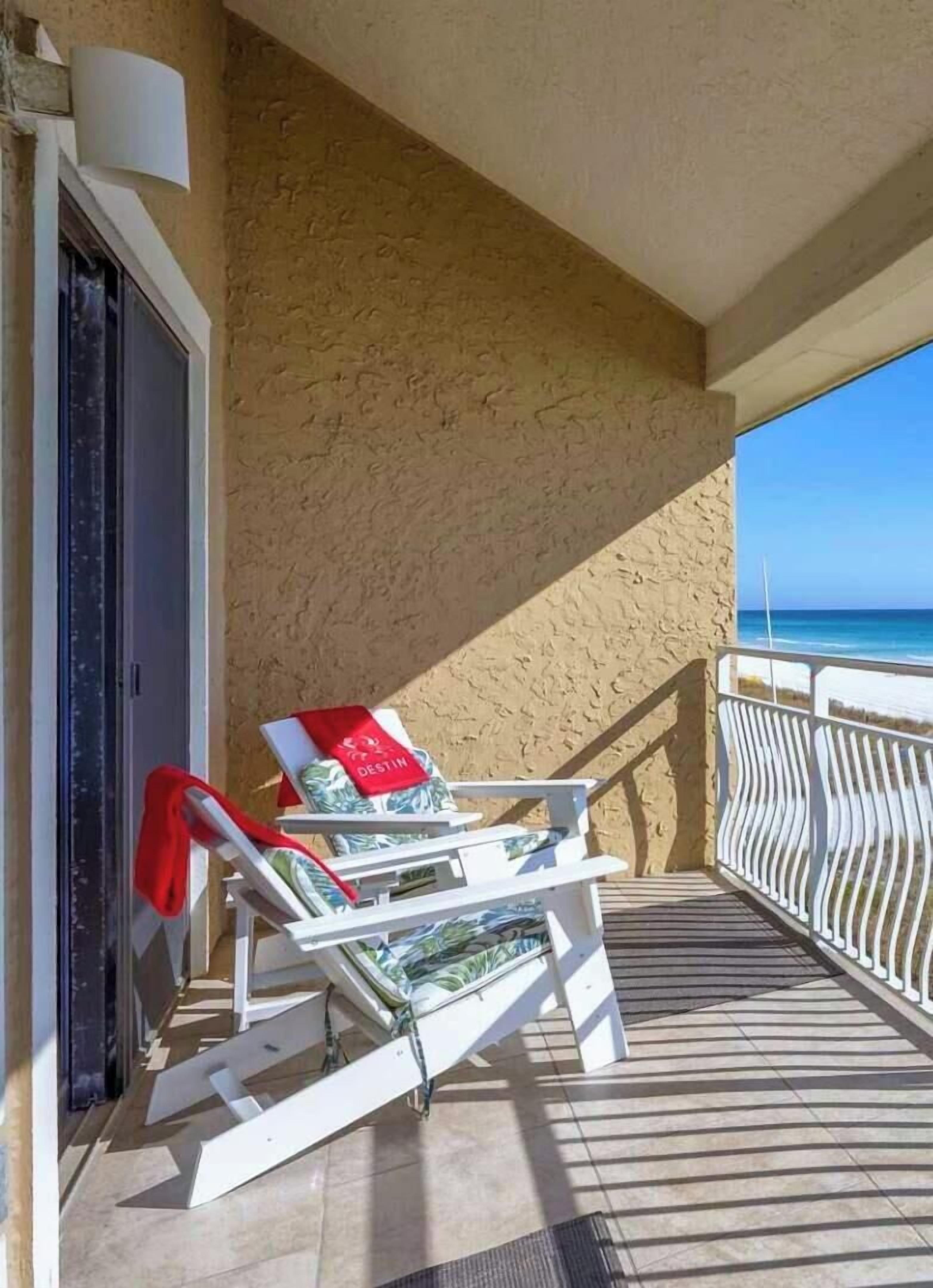 A balcony with a view of the beach and ocean, a chair, and a suitcase.