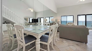 A spacious living area with a glass dining table, white chairs, a gray sofa, and a large window offering a view of the ocean. - Gulf View Getaway at Crystal Villas (Destin)