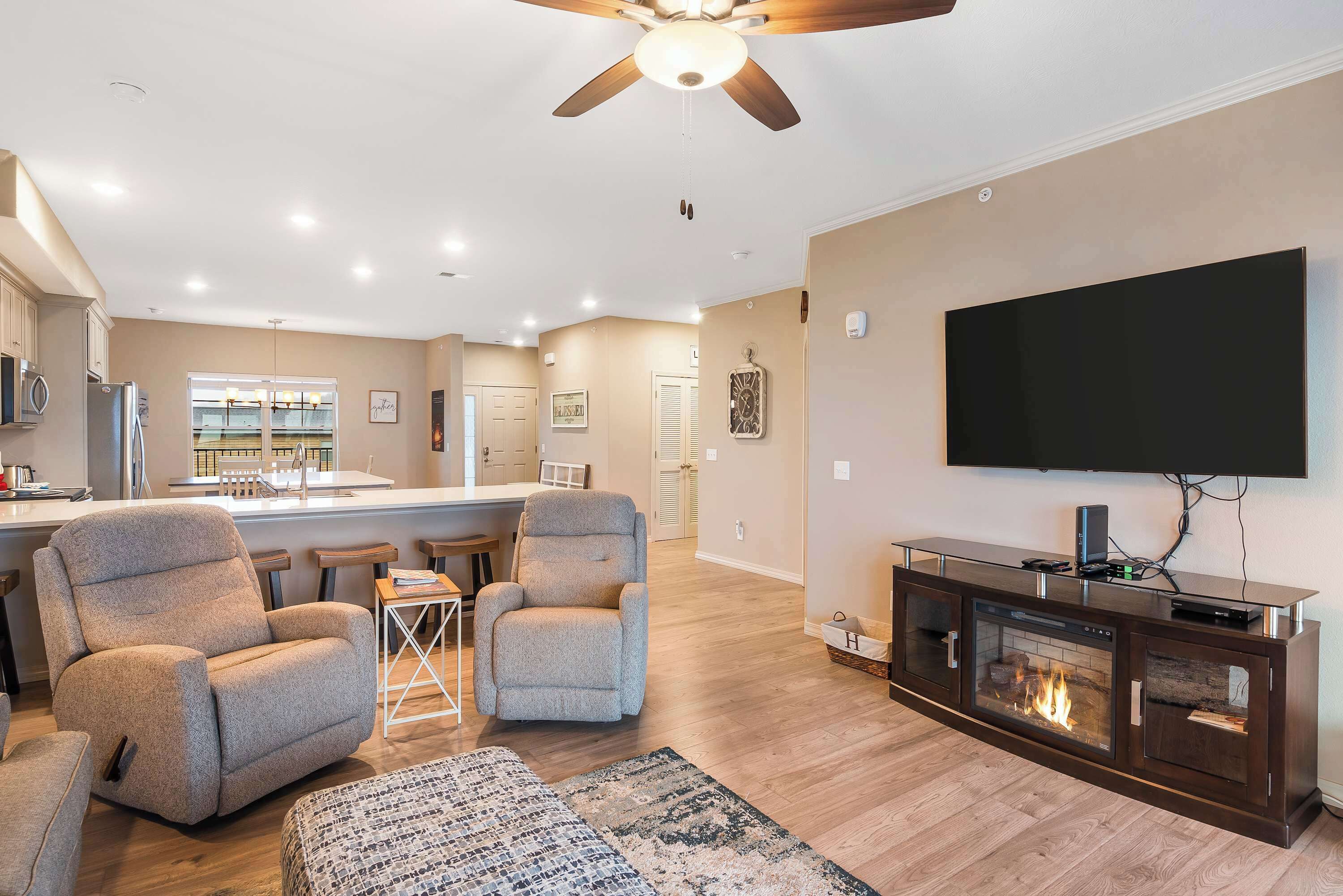 A modern living room with a fireplace, comfortable seating, a large TV, and a kitchen in the background.