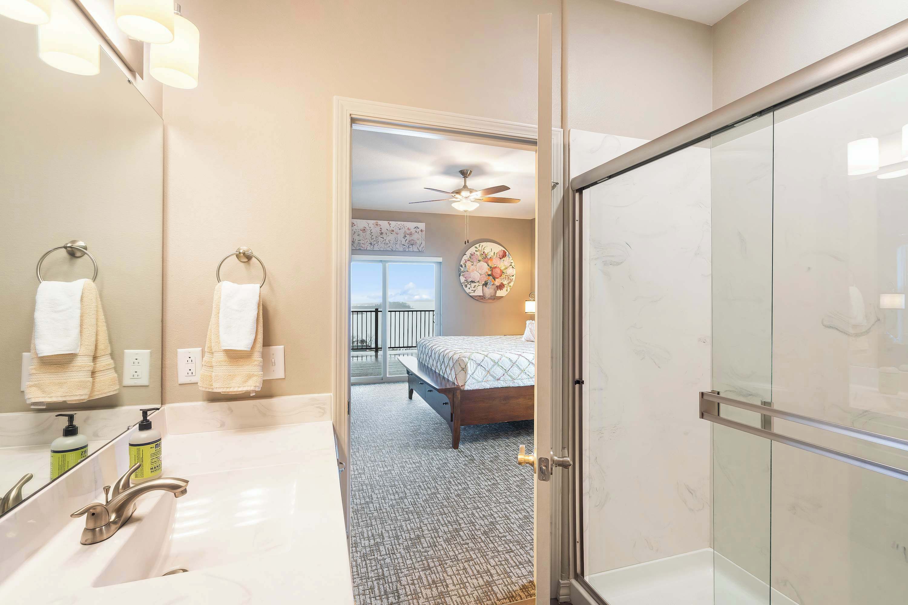 A bathroom with dual sinks, a shower, and a bed visible through an open door.