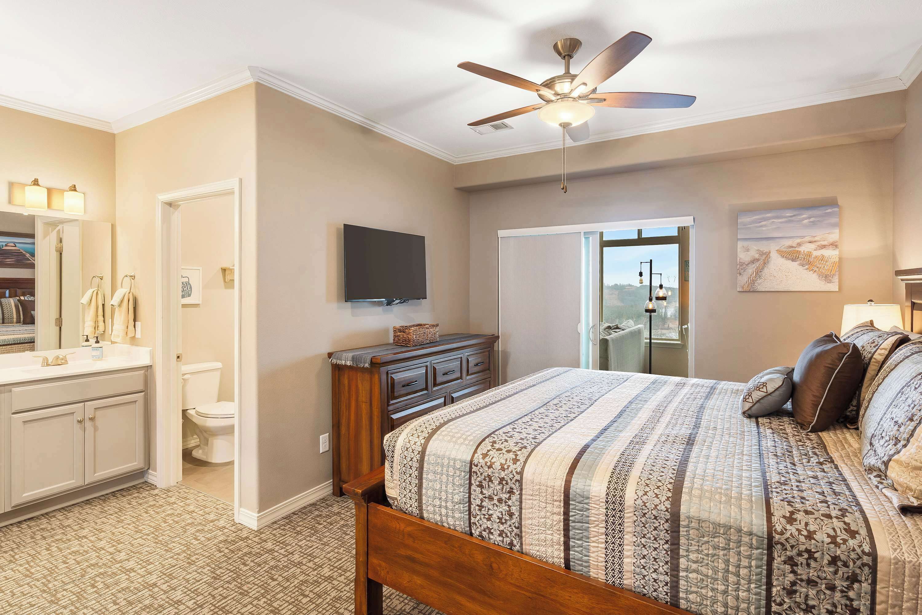 A bedroom with a bed, a ceiling fan, a dresser, a television, and a bathroom with a toilet and sink.