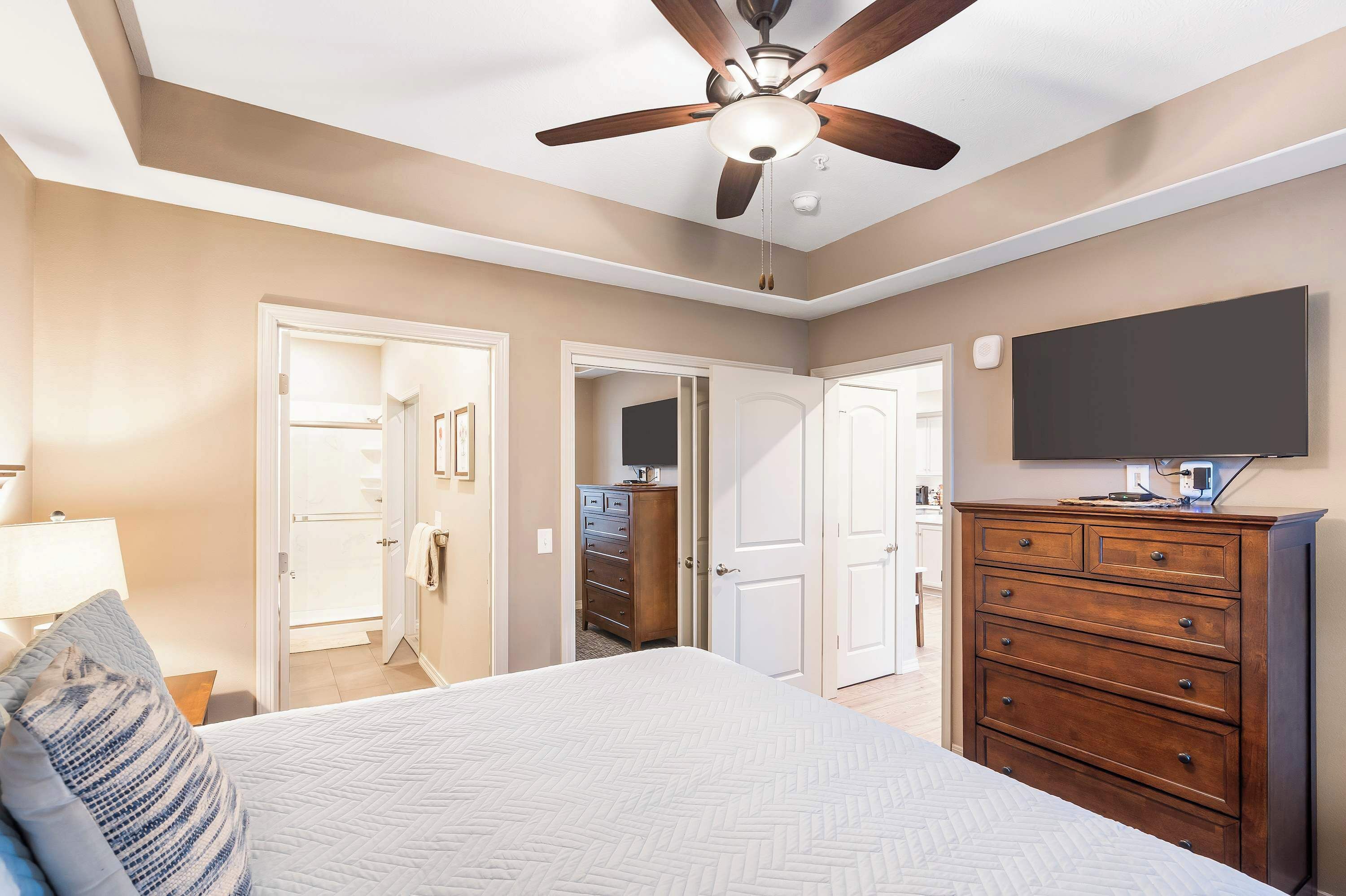 A bedroom with a bed, a dresser, a ceiling fan, and a TV on the wall.
