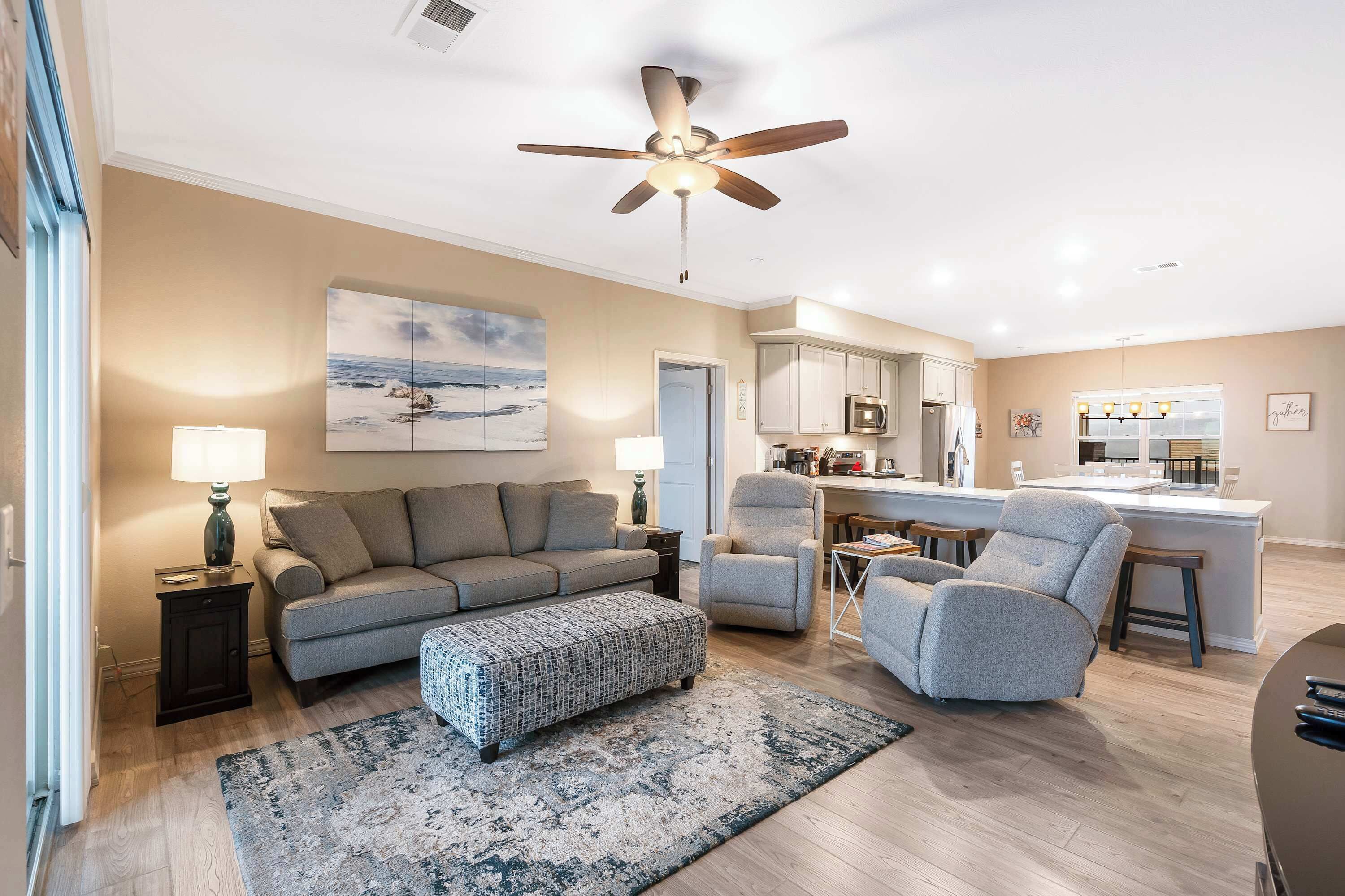 A modern living room with a sofa, ottoman, and armchairs, a ceiling fan, and a kitchen area with a bar.