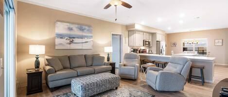 A modern living room with a sofa, ottoman, and armchairs, a ceiling fan, and a kitchen area with a bar.