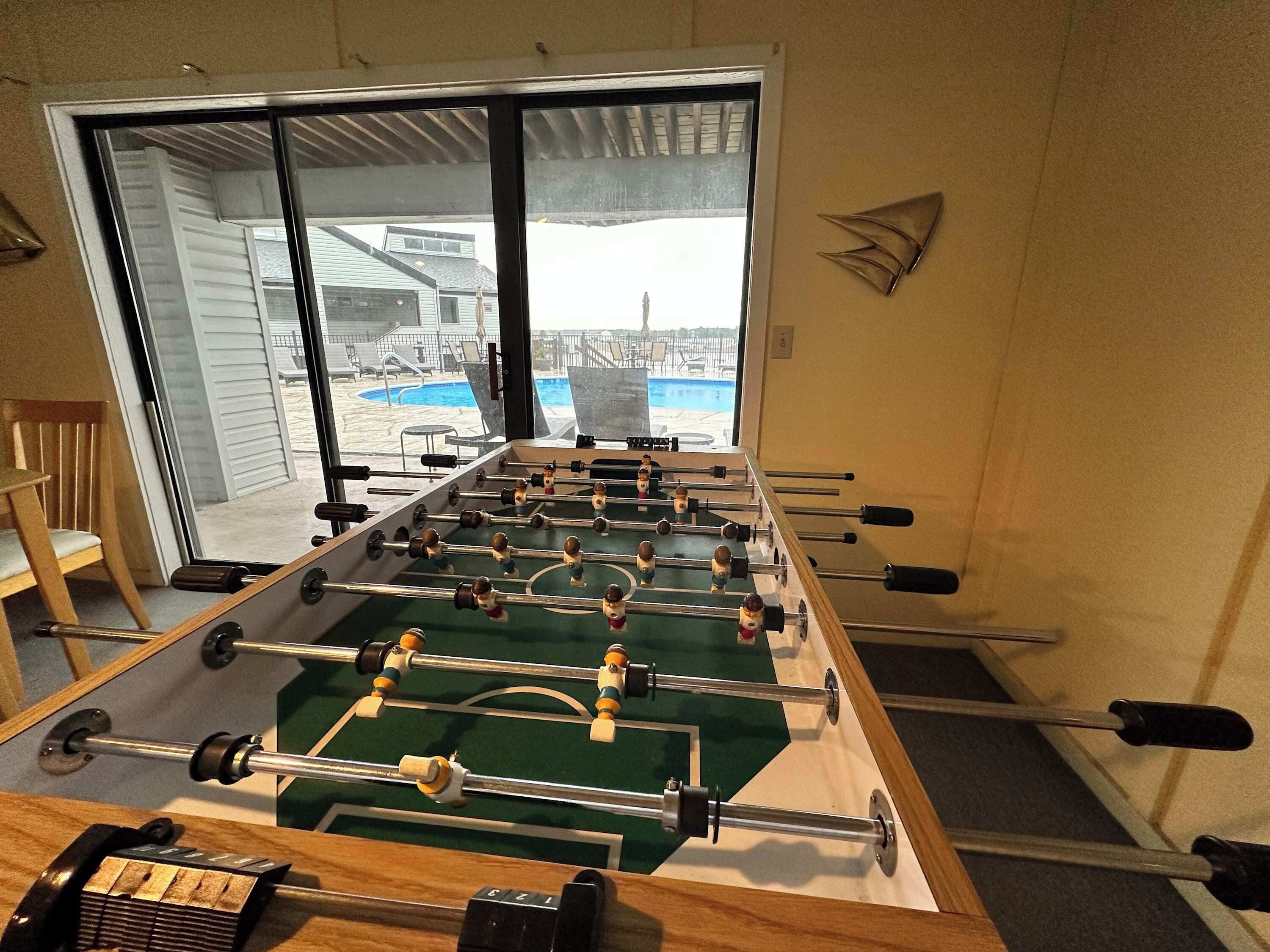 Una mesa de fútbol sobre una alfombra en una habitación con un amplio ventanal que da a una piscina.