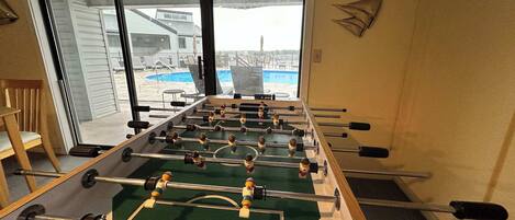Una mesa de fútbol sobre una alfombra en una habitación con un amplio ventanal que da a una piscina.