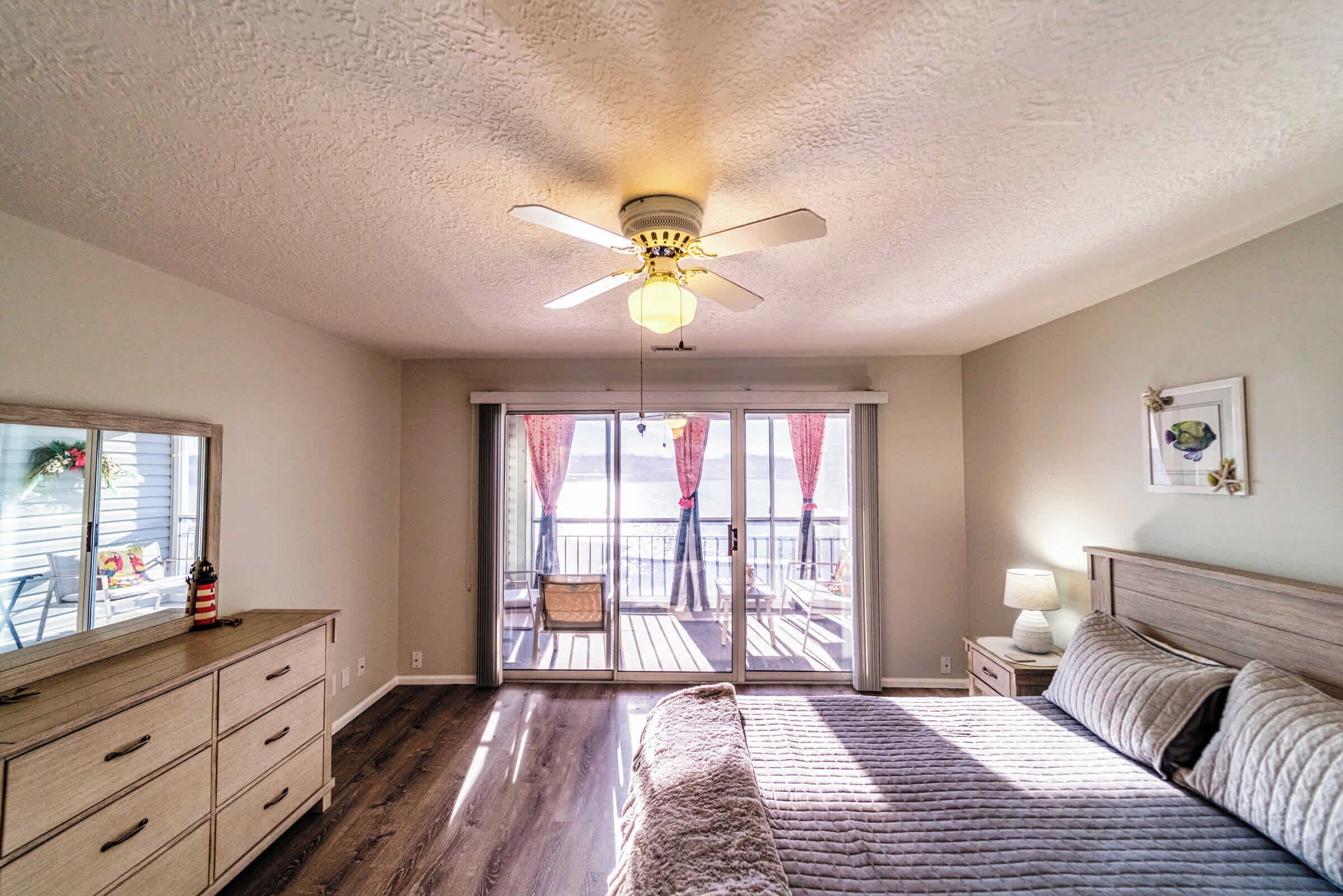 Une chambre à coucher comprenant un lit, une commode, une table de chevet, un ventilateur de plafond et une vue sur un balcon aménagé avec du mobilier d’extérieur.