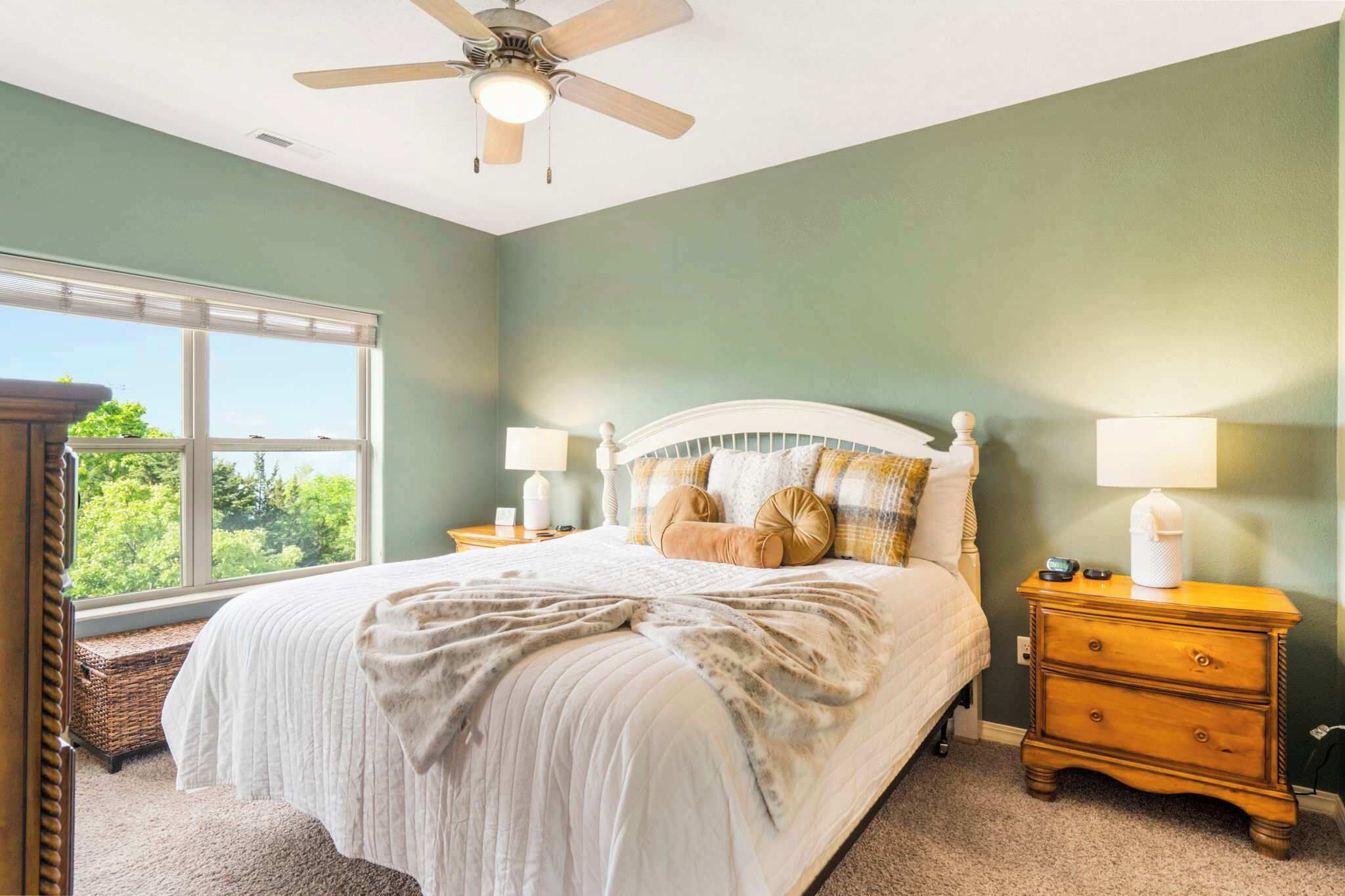 A bedroom with a bed, nightstands, a ceiling fan, and a window with a view of trees.