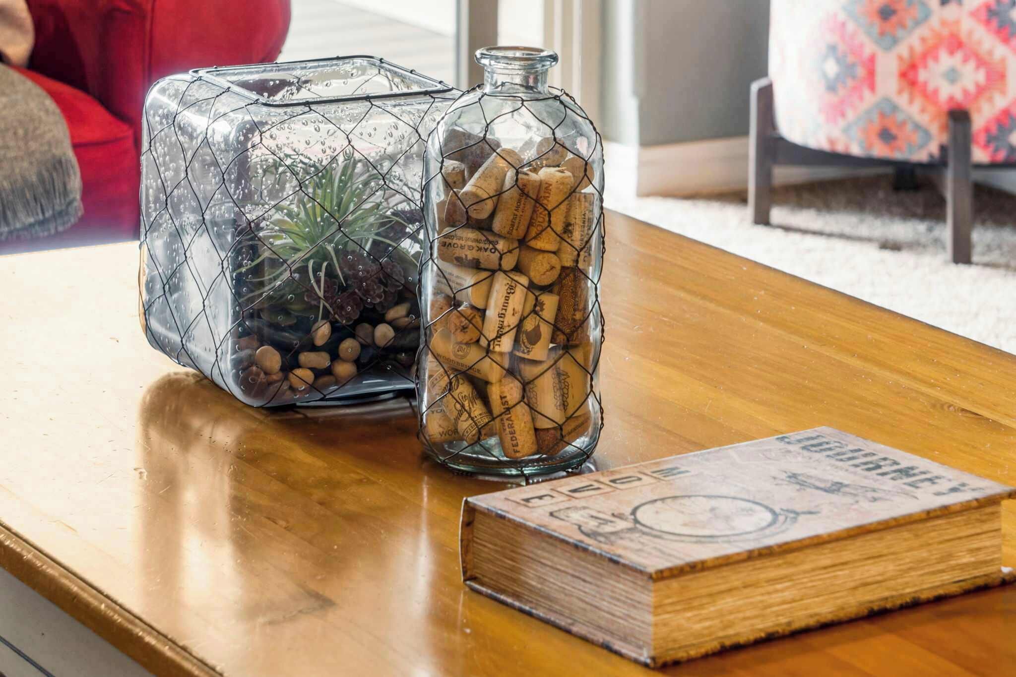 Two glass containers with wire frames, one filled with corks and the other with pebbles and a plant, on a wooden table with a book beside them.