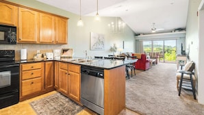 A modern kitchen with wooden cabinets, a dishwasher, and a large island. There is a dining area with a red sofa and a view of the outdoors. - Sunset Daydream Elegant Condo on Table Rock! (Branson)
