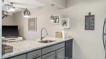 Una cocina moderna con encimeras de granito, un lavamanos de acero inoxidable y lavavajillas integrado. Hay lámparas colgantes, un reloj de pared y un letrero de bienvenida.
