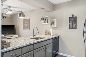 A modern kitchen with granite countertops, a stainless steel sink, and a built-in dishwasher. There are pendant lights, a wall-mounted clock, and a welcome sign. - Honey Hill - Pet Friendly - Indoor Pool (Branson)