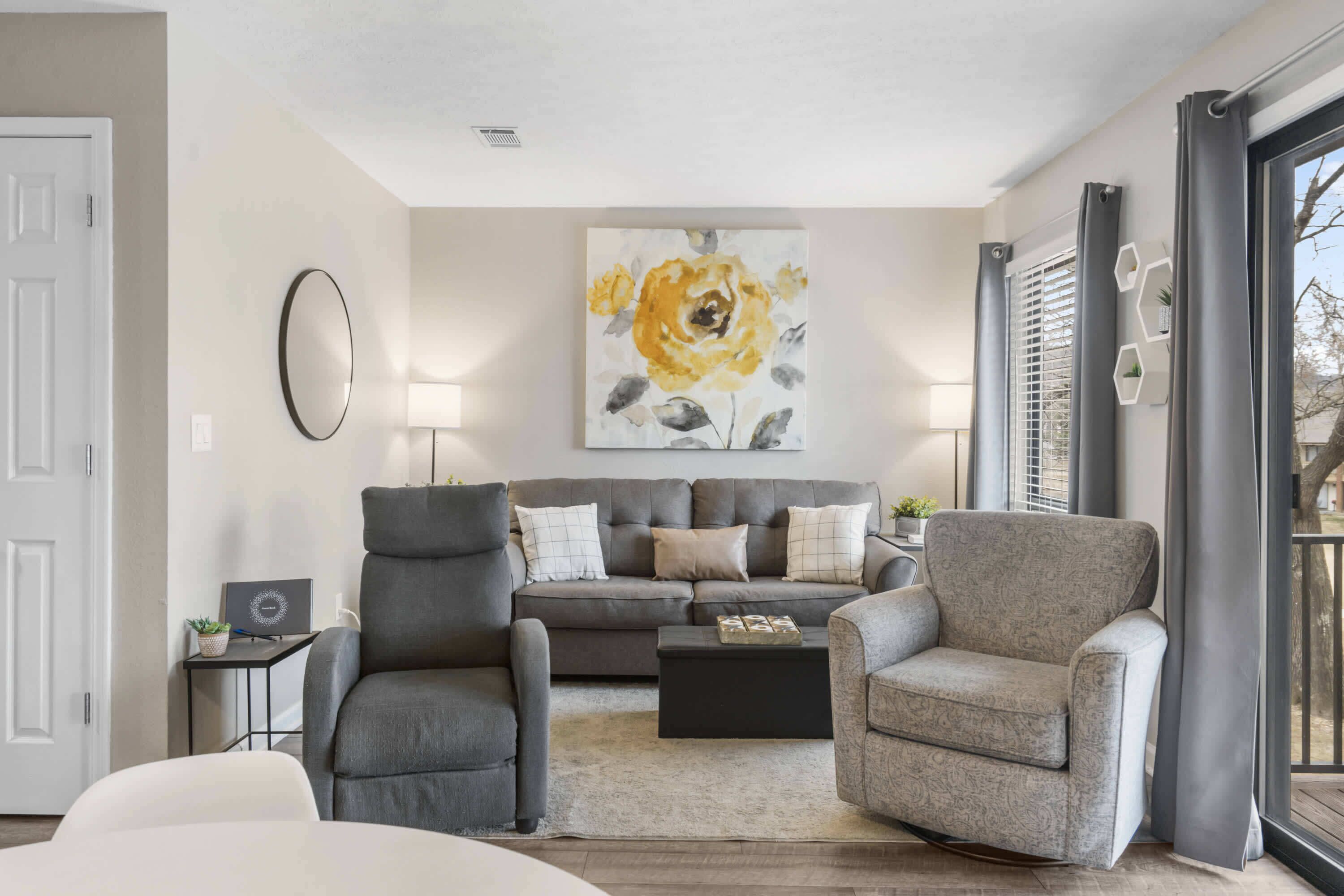 Un salon avec un canapé gris, deux fauteuils, une table basse et un grand tableau floral accroché au mur.
