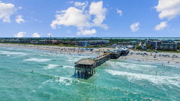 A beach with a pier extending into the ocean, surrounded by buildings and a clear sky.