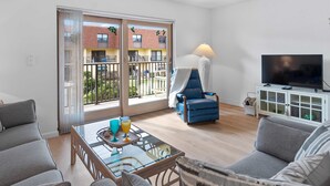 A modern living room with a glass table, two sofas, a blue armchair, a TV, and a balcony with a view of buildings and greenery.
