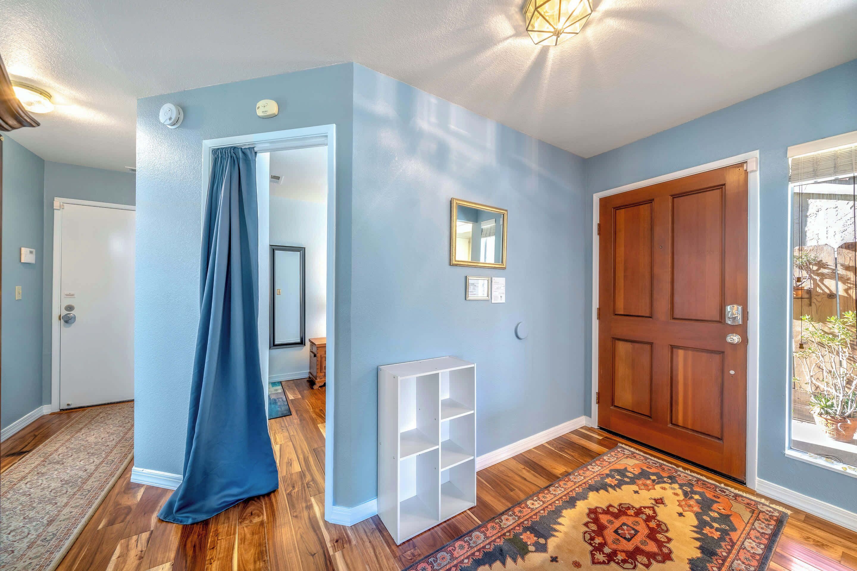 Un pasillo con pared azul, piso de madera, alfombra estampada, un mueble de almacenamiento blanco y una puerta con vista al exterior.