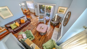 A spacious living room with a brown leather sofa, green armchairs, and a patterned ottoman. A dining area with a wooden table and chairs is visible through a sliding glass door leading to a patio with a view of a garden. - Succulents & Seashells Coastal Paradise by Beach (Marina)