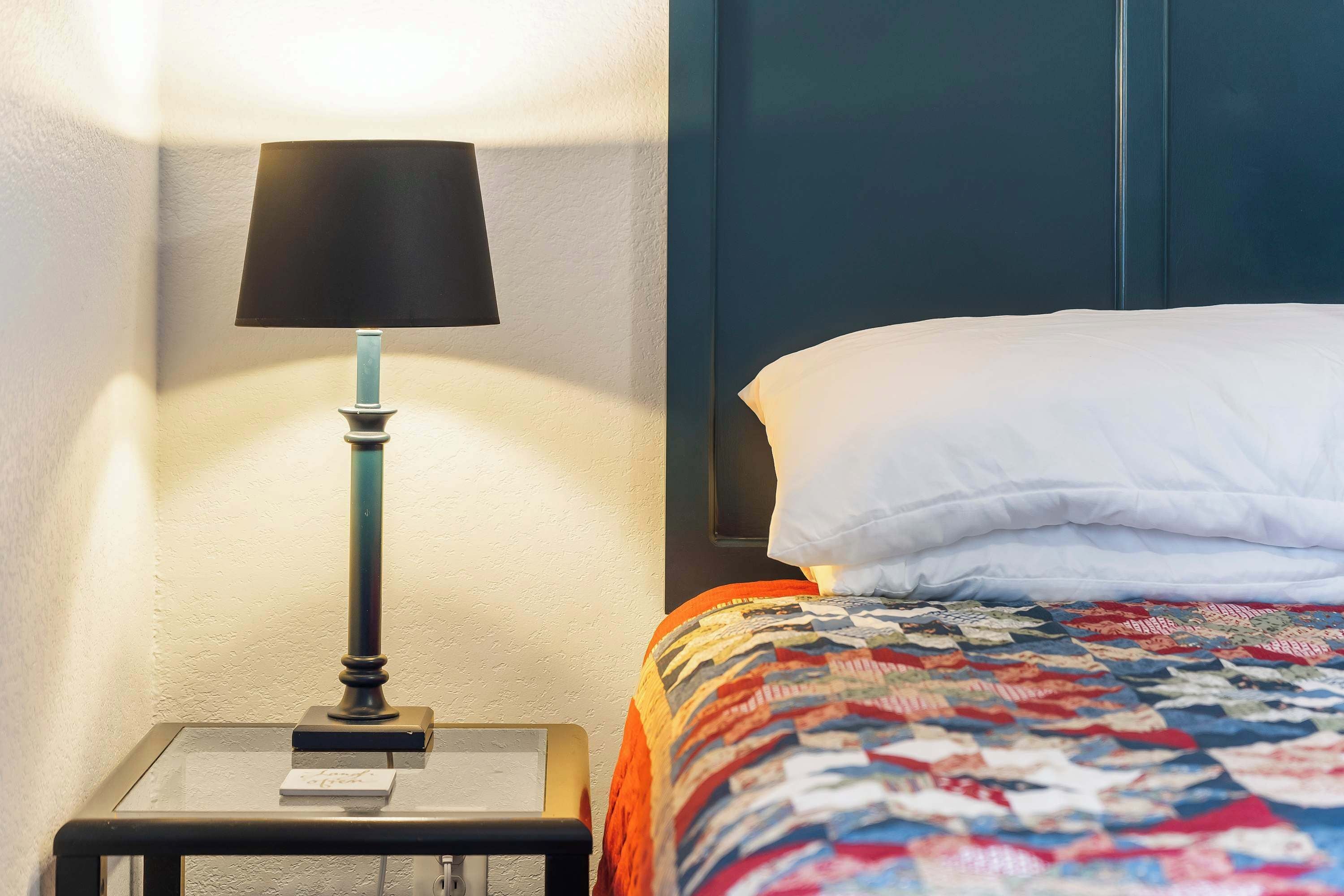 A neatly made bed with a colorful quilt, a black lamp on a bedside table, and a white wall.