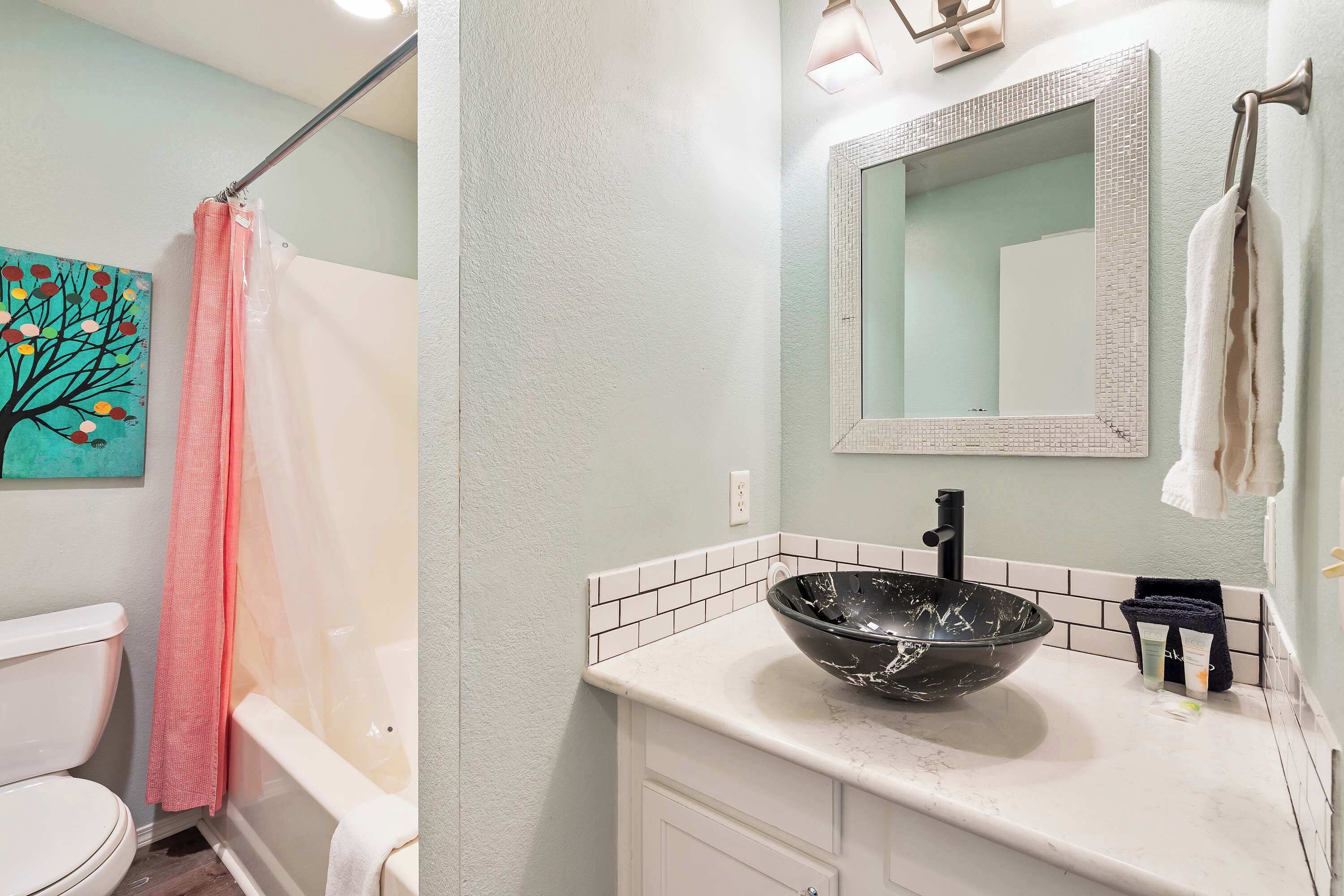 A bathroom with a white toilet, a black vessel sink, a white bathtub, a pink shower curtain, and a painting of a tree on the wall.