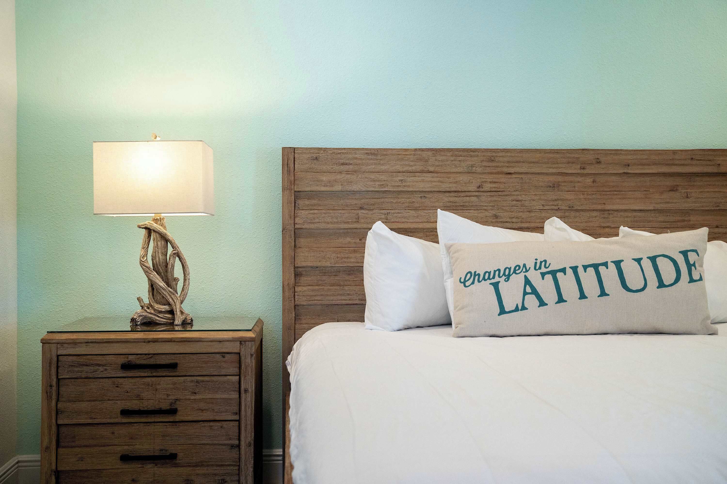 A neatly made bed with a pillow that reads "Changes in LATITUDE" and a wooden nightstand with a lamp beside it.
