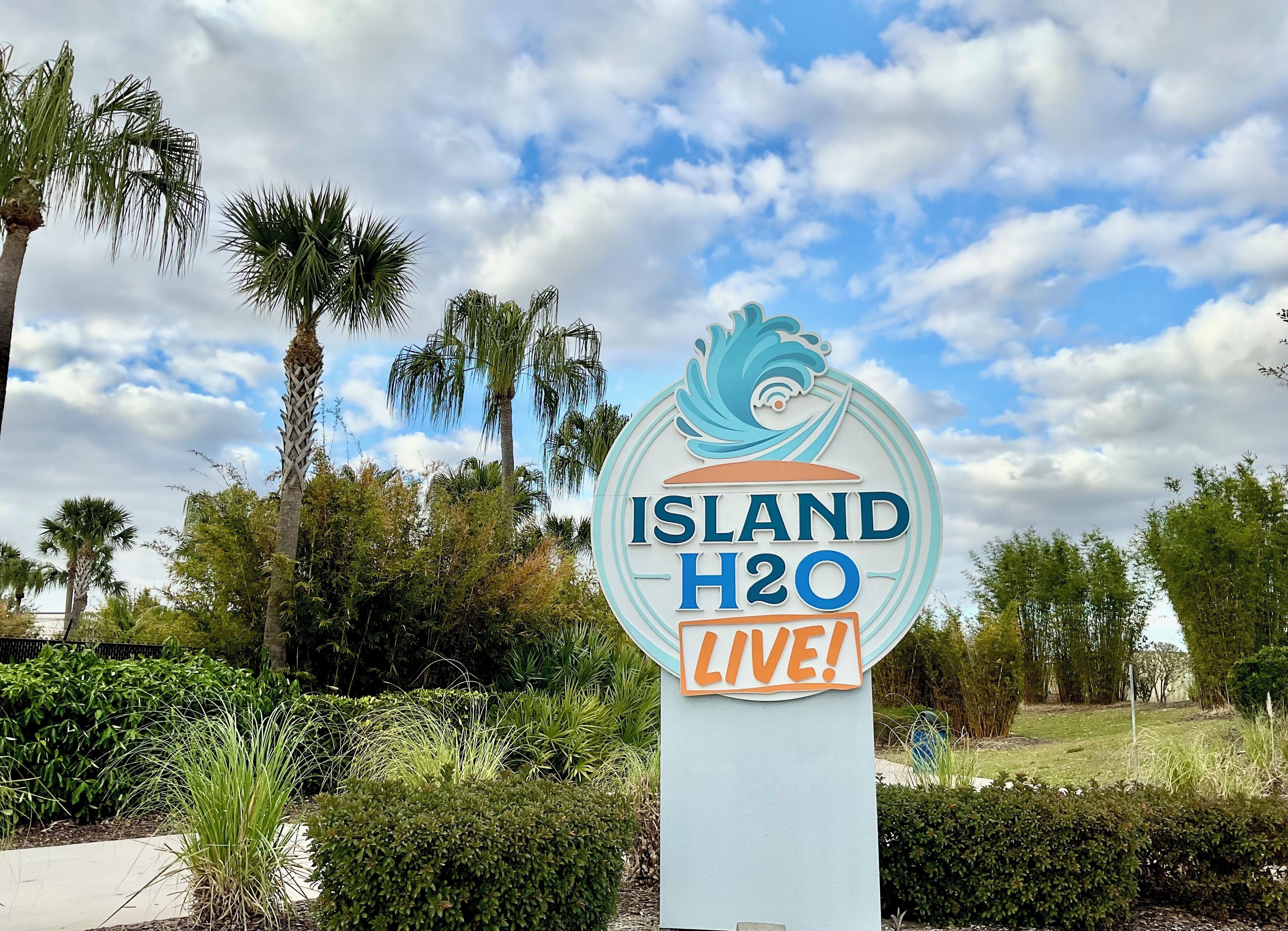 Un letrero con el texto 'ISLAND H2O LIVE!', rodeado de follaje y palmeras.